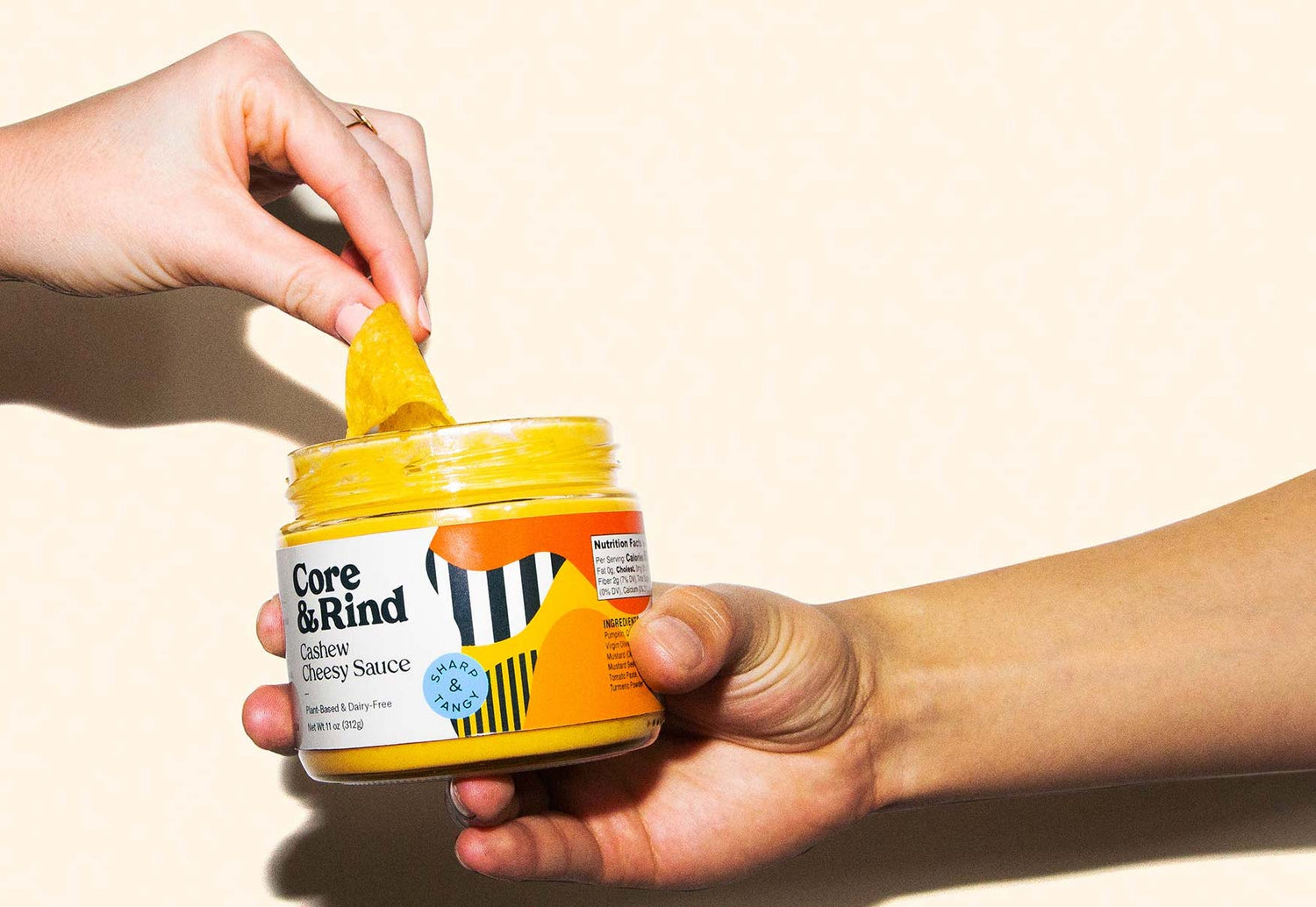 One hand holding a jar of cheesy sauce while another hand dips a chip into the jar